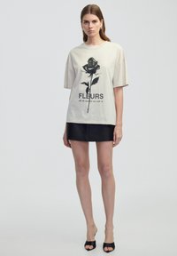 Béžové grafické tričko s potiskem černé růže a textem "FLEURS" v kombinaci s černou mini sukni. Materiál vypadá jemně a má uvolněný střih.