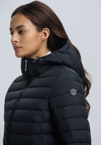 Frau mit langen braunen Haaren, die eine schwarze gesteppte Kapuzenjacke mit Reißverschluss und einem kleinen runden Logo-Patch am Ärmel trägt.