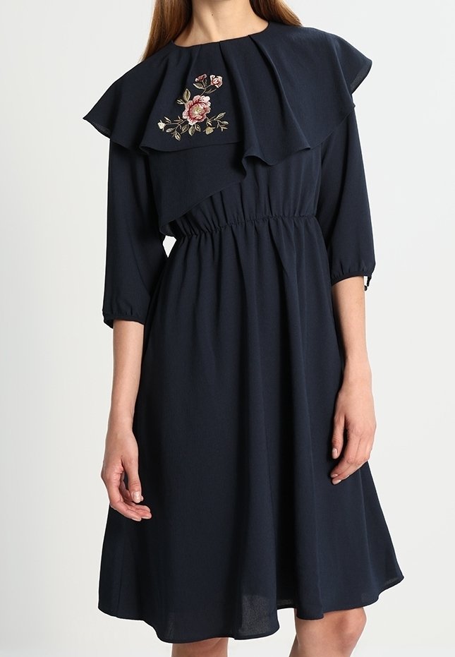 Femme portant une robe bleu marine longue jusqu'aux genoux avec des manches trois-quarts, un large col à couches, et une broderie florale sur la poitrine.