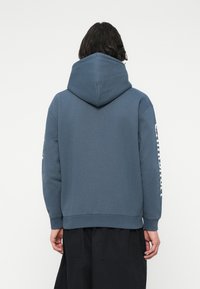 Plava hoodie od mekog materijala s velikom kapuljačom, rebrastim rukavima i logotipom ispisanim vertikalno na lijevom rukavu. Glatka tekstura.