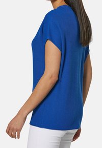 Niebieska, prążkowana bluzka z krótkim rękawem o luźnym kroju, okrągłym dekolcie i bocznymi rozporkami, pokazana w połączeniu z białymi spodniami.