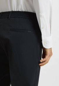 Navy trousers with an elastic waistband; featuring a rear pocket and textured fabric. Complemented by a white, buttoned long-sleeve shirt.