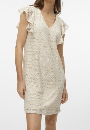 Femme portant une robe en dentelle couleur crème avec des manches volantées et un motif en zigzag, debout devant un fond uni.