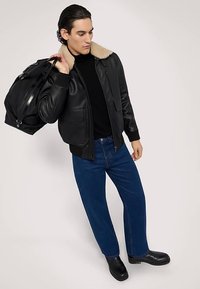 Černá kožená bomber bunda s béžovým fleecovým límcem, tmavý rolák, modré rovné džíny a černé boty, držící černou tašku.