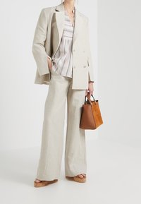 Beige Leinenanzug mit weit geschnittenen Hosen, kombiniert mit einer gestreiften Bluse in Pastellfarben. Eine braune Wildlederhandtasche und Platform-Sandalen vervollständigen den Look.