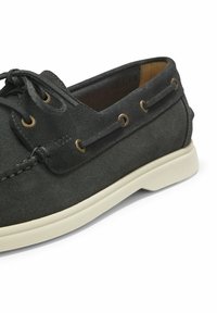 Chaussure bateau en daim gris foncé avec semelle en caoutchouc blanche, à lacets, avec des lacets en cuir passant à travers des œillets métalliques le long des côtés.