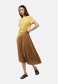 T-shirt en coton jaune associé à un pantalon large en brown. La tenue est complétée par des sandales vert foncé au design entrelacé.