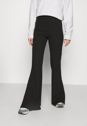 Persona que lleva pantalones negros acampanados de talle alto, camiseta gris de manga larga y zapatillas blancas y verdes, de pie frente a un fondo liso.