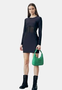 Zwarte mini-jurk met lange mouwen en een netpaneel bij de taille, gecombineerd met een groene handtas en zwarte veterschoenen tot de enkel.