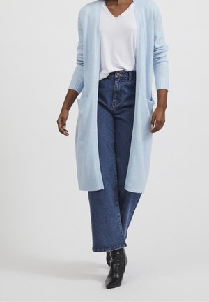 Frau trägt hellblauen langen Cardigan, weißes V-Ausschnitt-Shirt, blaue Jeans und schwarze Stiefeletten, steht vor einfarbigem Hintergrund.