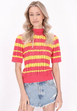 Vrouw met lang blond haar draagt een kort mouw gebreide trui met roze en gele strepen en lichtblauwe beschadigde denim shorts.