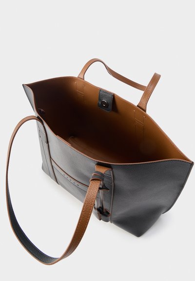 Ανοιχτή τσάντα τύπου τσάντα ώμου από μαύρο και καφέ δέρμα με μακριές λαβές, που δείχνει το λείο εσωτερικό και το μαγνητικό κούμπωμα σε ουδέτερο φόντο.