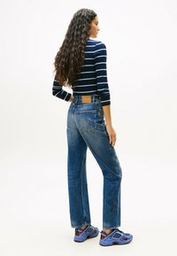 Mujer con cabello rizado y largo, vistiendo una camiseta de rayas azul marino, jeans de tiro alto azul con bordado de flores y zapatillas deportivas moradas, de pie de lado.