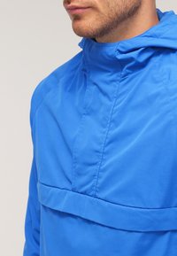 Blå huva jacka med en texturerad finish, utrustad med en framficka och en hög krage. Lättviktsdesign som är lämplig för aktiv användning.