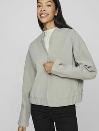 Šviesiai pilka zip-up striukė su minimaliu dizainu, turinti apvalų iškirpimą ir raštuotą apačią, pagaminta iš lygaus audinio, uždėta ant megztinio.