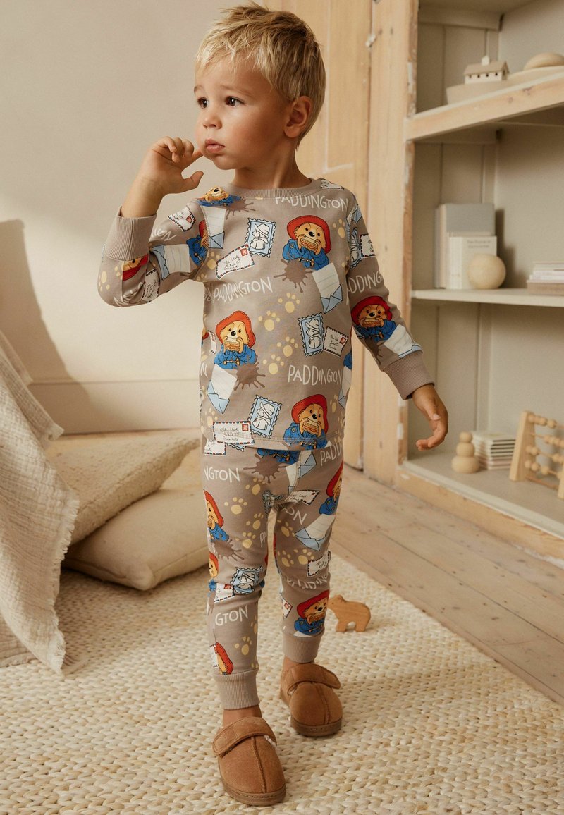 Unge barn i beige Paddington-inspireret pyjamas står barfodet på et vævet tæppe i et hyggeligt, blødt oplyst rum med trægulv.