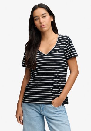 Kvinde med langt mørkt hår iført en sort-hvid stribet V-hals T-shirt og lyseblå jeans, stående med den ene hånd i lommen.