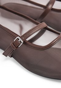Mocasines marrones con un empalme de malla transparente y una correa de cuero marrón. Presenta una hebilla de metal plateado para el ajuste y un diseño de punta redonda.