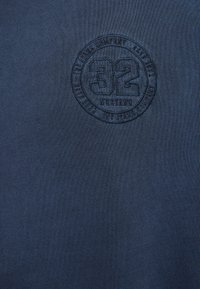Sweat-shirt bleu marine avec une texture côtelée et un logo circulaire embossé affichant le numéro "32" au centre.