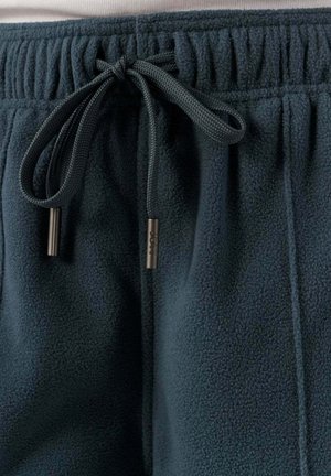 Joggers de felpa en un tono verde oscuro con una cinturilla elástica y un cordón de ajuste. Presenta puntas de metal en el cordón. Textura suave, corte relajado.