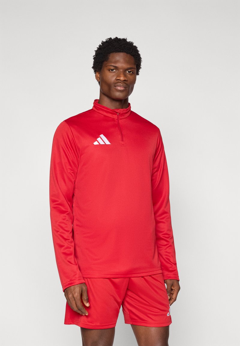 Κόκκινο αθλητικό μπουφάν με ημίψηλο γιακά και μακριά μανίκια, κατασκευασμένο από λείο πολυεστέρα. Διαθέτει λευκό λογότυπο στο αριστερό στήθος και συνδυάζεται με σορτς.