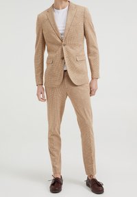 Ljust brun kostym i texturerat tyg; kavaj med två framfickor och en knapp; slim-fit byxor; ihop med mörkbruna skor.