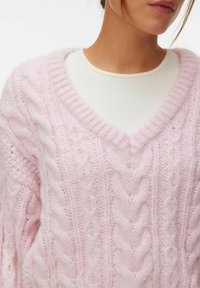 Femme portant un pull en maille torsadée à col en V, duveteux et rose pastel, par-dessus un haut blanc côtelé, vue de la poitrine avec les cheveux partiellement visibles.