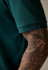Polo de manga corta verde oscuro con una tela texturizada, que presenta un cuello doblado. Un tatuaje floral detallado visible en el antebrazo.