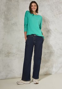 Maglietta azzurra a maniche lunghe con dettagli a costine, abbinata a jeans in denim a gamba larga blu scuro e sneakers beige su uno sfondo testurizzato.