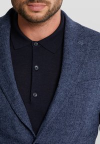 Marinblå herringbone-blazer med en texturerad yta, lagd över en mörk polotröja med knapplist och krage.