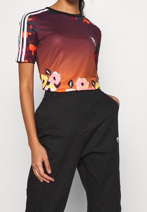 Personne portant une chemise à manches courtes avec un dégradé floral et des bandes blanches sur les manches, ainsi qu'un pantalon cargo noir, main dans la poche.