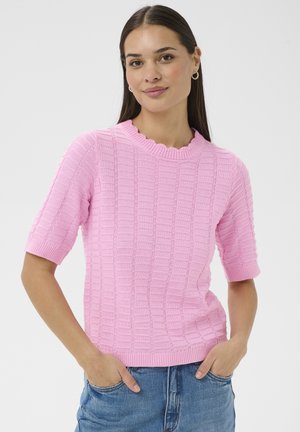 Vrouw met lang donker haar, draagend grote creolen, een roze textuur kort mouw trui en een blauwe spijkerbroek, met handen in de zakken, staand op een witte achtergrond.