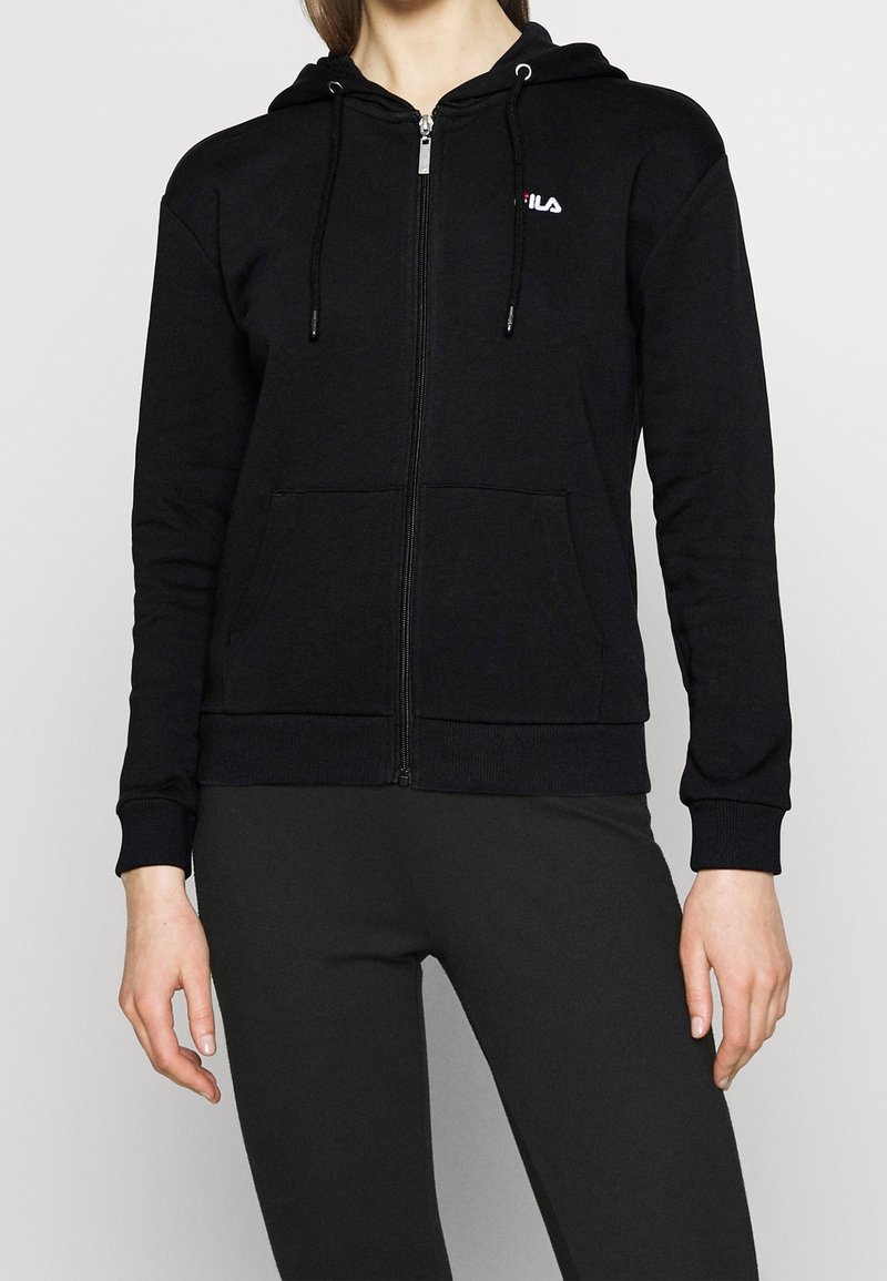 Sudadera negra con cremallera fabricada en tela suave, con un bolsillo frontal, cordones ajustables y un pequeño logo blanco en el lado izquierdo del pecho.