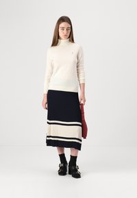 Un pull col roulé crème, une jupe plissée marine avec des rayures crème et marine, des sandales à talons noires, tenant un sac rouge. Texture lisse, design ajusté.