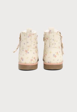Par de botines color blanco roto con pequeño estampado floral en tonos pastel, suelas marrones y correas laterales, mostrados desde atrás sobre un fondo liso.