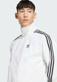Vit zip-up jacka med hög krage, prydd med svart Adidas-logotyp och tre svarta ränder längs axlarna. Lättviktig tyg med slät textur.