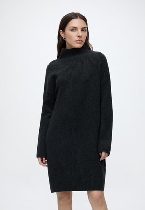 Frau trägt ein langes schwarzes, geripptes Strickkleid mit hohem Kragen und steht vor einem weißen Hintergrund.
