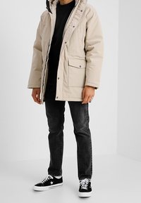 Parka beige con capucha, cremallera frontal y botones de presión. Cuenta con un gran bolsillo en el pecho izquierdo y jeans negros combinados con zapatillas negras.
