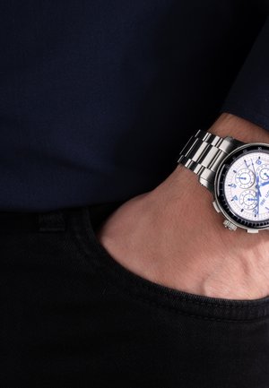 Linkerhand in de zak van een zwarte spijkerbroek, met een zilveren metalen chronograafhorloge met wijzerplaat in wit en blauwe wijzers, tegen een achtergrond van een donkerblauw overhemd.