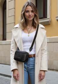 Petit sac en cuir noir texturé porté en bandoulière, avec une fermeture éclair et des accents dorés, associé à un manteau crème sur un haut blanc et un jean bleu.