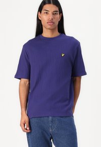 Camiseta de manga corta morada hecha de tela texturizada. Presenta un pequeño logo amarillo bordado en el lado izquierdo del pecho. Ajuste casual.