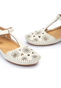 Sandales en cuir blanc avec des motifs floraux découpés, bout rond, bride à la cheville et couture tressée le long du bord. Semelle plate.