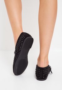 Czarne zamszowe buty wsuwane z ozdobnymi ćwiekami, płaską gumową podeszwą i dekoracyjną kokardą po boku.