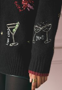 Suéter negro con diseños de cócteles bordados, acentos de lentejuelas y un dobladillo rojo acanalado. El texto dice "Extra Orty" y "Salut!"