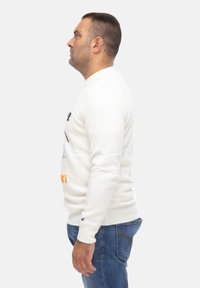 Maglione bianco lavorato a maglia con collo rotondo, con testo blu e arancione sul davanti, abbinato a jeans di denim blu. Vista laterale.