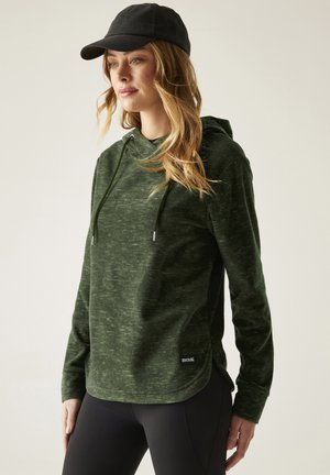 Donna che indossa un pullover con cappuccio verde scuro e un cappellino da baseball nero, in piedi con le mani lungo i fianchi su sfondo semplice.