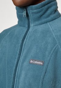 Veste en polaire avec col montant, fermeture éclair à l'avant, de couleur teal. Logo Columbia sur la poitrine. Texture douce avec coutures surpiquées.