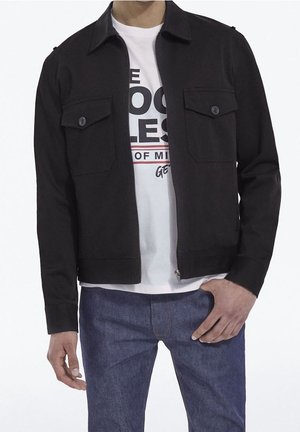 Homme portant une veste noire zippée avec deux poches avant à boutons sur un t-shirt graphique blanc et un jean bleu, debout sur fond blanc.