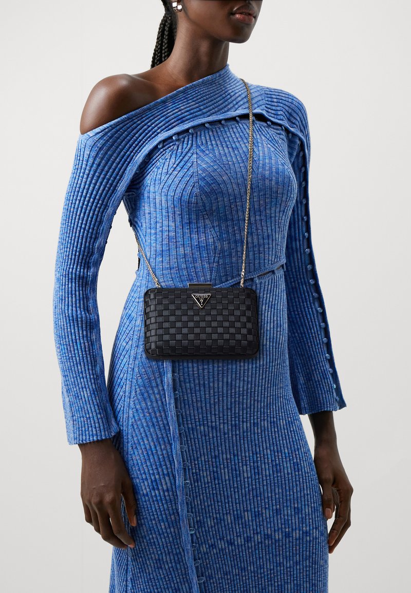 Borsa a tracolla intrecciata nera con tracolla in catena argentata, indossata sopra un vestito blu a coste con texture, caratterizzato da un design off-the-shoulder e dettagli con bottoni.