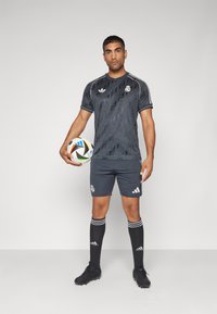 Camiseta atlética gris con un diseño estampado, pantalones cortos a juego, calcetas negras con rayas blancas y botas negras, sosteniendo un balón de fútbol.